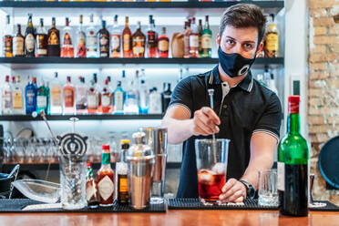 Seriöser männlicher Barkeeper, der einen Cocktail mit alkoholischen Getränken und Eiswürfeln zubereitet, während er am Tresen einer Bar steht - ADSF16646