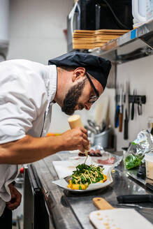 Side view of busy male chef in uniform adding sauce on top of dish with greenery while working in kitchen of cafe - ADSF16650