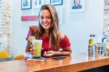 Lächelnder weiblicher Kunde sitzt am Tresen eines Cafés und genießt ein süßes Entgiftungsgetränk mit Kiwi, während er in die Kamera schaut - ADSF16653