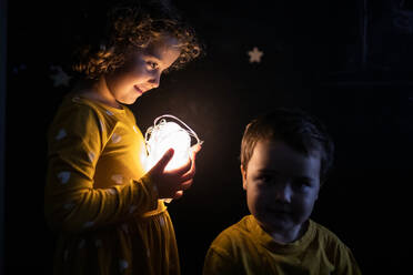 Side view of curious siblings in pajamas standing in dark room with illuminated garland - ADSF16658