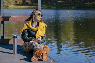 Smiling woman having coffee while relaxing pier over lake during weekend - KNTF05747