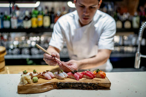 Male chef garnishing sushi on wooden serving tray in kitchen at restaurant - OCMF01764