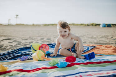 Niedliche kleine Baby spielen, während auf bunten Handtuch am Strand bei Sonnenuntergang sitzen - GMLF00724