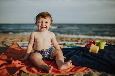 Nettes männliches Kleinkind lächelnd am Strand sitzend an einem sonnigen Tag - GMLF00730