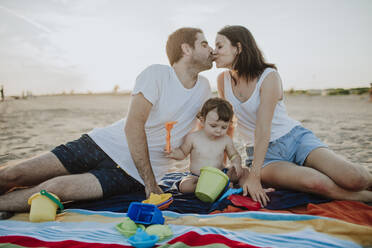 Ehemann und Ehefrau küssen sich, während ihr Sohn am Strand mit Spielzeug spielt - GMLF00736
