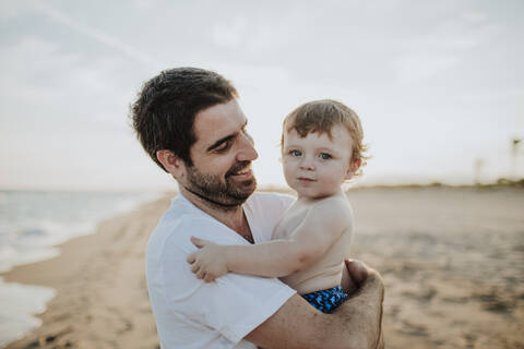 Smiling father carrying baby boy at beach during sunset stock photo