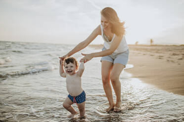 Fröhliche Mutter und Sohn genießen im Wasser am Strand an einem sonnigen Tag während des Sonnenuntergangs - GMLF00743