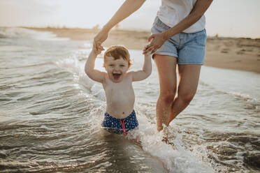 Midsection der Mutter mit fröhlichen Baby Sohn genießen im Wasser am Strand an einem sonnigen Tag während des Sonnenuntergangs - GMLF00744