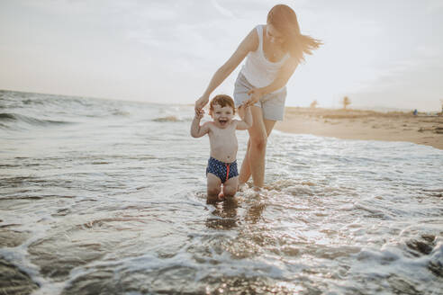 Mother and son enjoying in water at beach on sunny day during sunset - GMLF00746