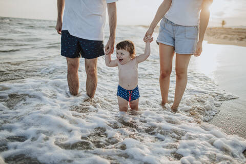 Midsection of mother and father with son standing in water at beach during sunset stock photo