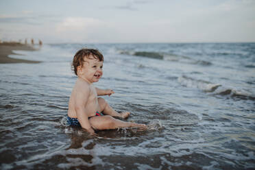 Male toddler enjoying while sitting in water at beach during sunset - GMLF00749