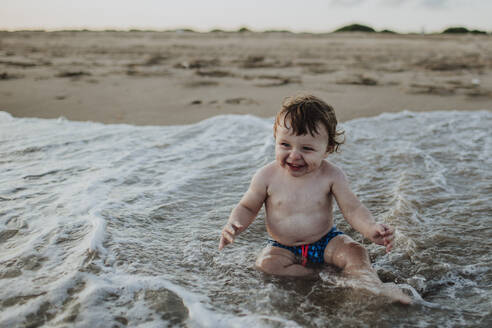 Fröhlicher kleiner Junge spielt im Wasser am Strand bei Sonnenuntergang - GMLF00750