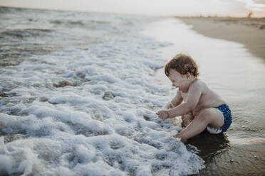 Cute little boy laughing while sitting in water at beach during sunset - GMLF00753