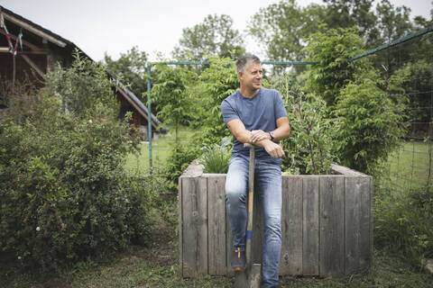 Smiling handsome man holding spade looking away while leaning on wooden raised bed at vegetable garden stock photo