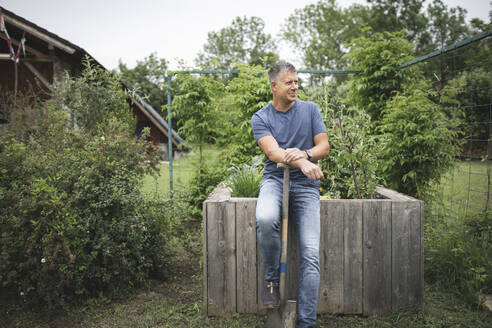 Smiling handsome man holding spade looking away while leaning on wooden raised bed at vegetable garden - HMEF01078