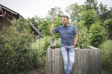 Gut aussehender Mann, der einen Spaten hält und wegschaut, während er sich auf ein hölzernes Hochbeet im Gemüsegarten stützt - HMEF01079