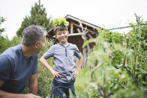 Verspielter Junge mit Pflanze auf dem Kopf schaut Vater im Garten an, lizenzfreies Stockfoto