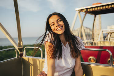 Cheerful young woman enjoying Ferris wheel ride against sky at sunset - OYF00218
