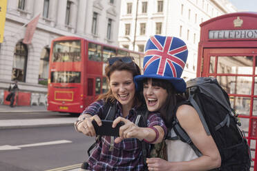 Glückliche Frau nimmt Selfie mit Freund trägt britische Flagge Hut gegen rote Telefonzelle in der Stadt - AJOF00279