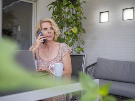 Nachdenkliche weibliche Fachkraft, die am Telefon spricht, während sie zu Hause mit einem Laptop arbeitet - LAF02497