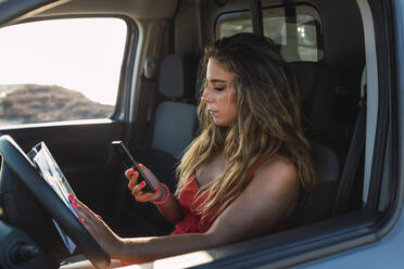 Junge Frau mit Landkarte, die ein Mobiltelefon benutzt, während sie im Wohnmobil am Strand sitzt - DCRF00960
