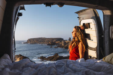 Young woman with hand in hair leaning on camper van door at beach - DCRF00967