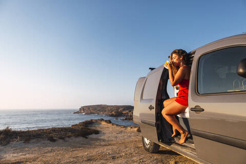 Woman drinking coffee while sitting in camper van at beach stock photo