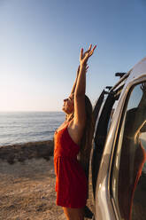 Frau mit erhobener Hand am Wohnmobil am Strand stehend - DCRF00969