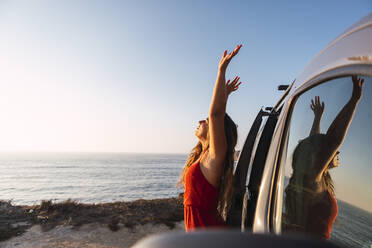Junge Frau mit erhobener Hand an einem Wohnmobil am Strand stehend - DCRF00971