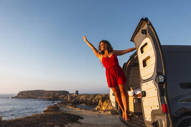 Lächelnde Frau mit erhobener Hand, die an der Tür eines Wohnmobils am Strand hängt - DCRF00981