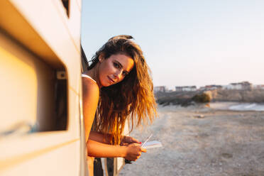 Frau mit Buch und Stift schaut aus der Tür eines Wohnmobils am Strand - DCRF00986