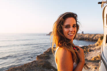 Smiling woman standing by camper van door at beach - DCRF00988