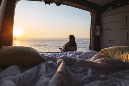 Frau bewundert die Aussicht auf den Sonnenuntergang, während sie auf einem Stuhl neben einem Wohnmobil am Strand sitzt - DCRF00992
