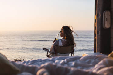 Young woman drinking white wine while sitting on chair by camper van at beach during sunset - DCRF00993