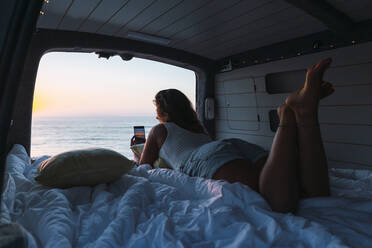 Woman taking photo of sunset through phone while lying in camper van at beach - DCRF00996