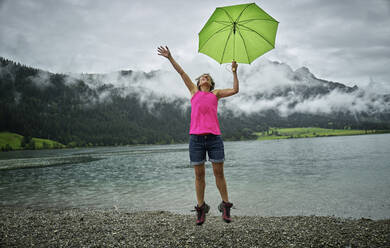 Glückliche reife Frau, die einen Regenschirm hält, während sie in der Regenzeit am Haldenseeufer springt - DIKF00534