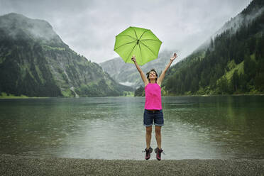 Glückliche reife Frau mit Regenschirm beim Springen am Vilsalpsee während der Regenzeit - DIKF00537