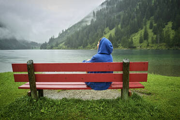 Frau mit blauer Kapuze sitzt auf einer Bank am Seeufer und schaut auf die Berge - DIKF00543