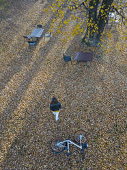 Drone view of woman walking towards chairs and tables at park during autumn - KNTF05753