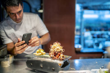 Chefkoch fotografiert Lebensmittel mit dem Handy, während er in einer Großküche steht - OCMF01770