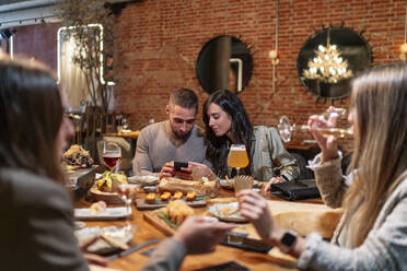 Frauen unterhalten sich beim Weintrinken mit Freunden und benutzen dabei ein Mobiltelefon im Hintergrund im Restaurant - OCMF01776