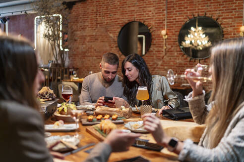 Frauen unterhalten sich beim Weintrinken mit Freunden und benutzen dabei ein Mobiltelefon im Hintergrund im Restaurant - OCMF01776