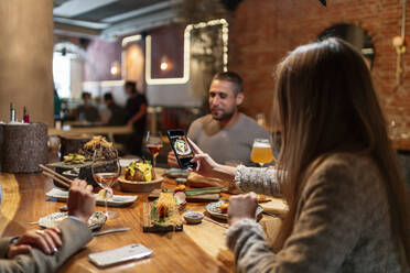 Junge Frau, die ihr Essen mit dem Handy fotografiert, während sie mit Freunden im Restaurant sitzt - OCMF01779