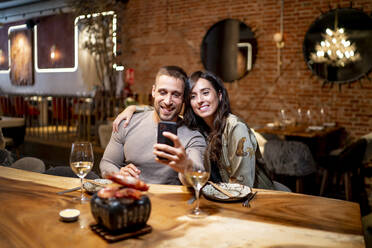 Smiling friends taking selfie while sitting by table at restaurant - OCMF01782