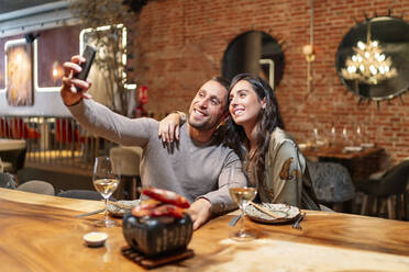 Smiling friends taking selfie through mobile phone while sitting by table at restaurant - OCMF01783