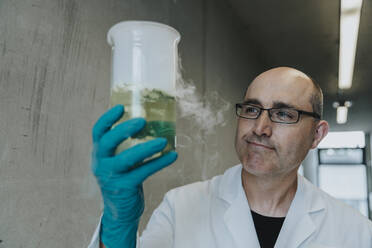 Scientist holding liquid flask with evaporating smoke while standing at clinic corridor - MFF06304