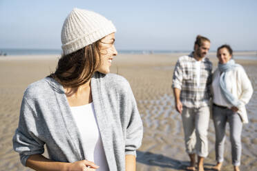 Junge Frau schaut über die Schulter zu einem Mann und einer Mutter, die im Hintergrund am Strand spazieren gehen - UUF21671