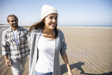 Young woman holding hand of man while walking on beach - UUF21677