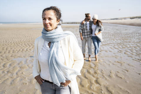 Woman with hands in pockets looking away while walking with couple in background at beach stock photo