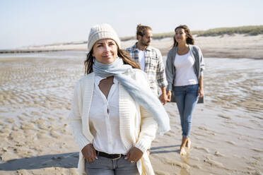 Smiling woman looking away while walking with couple holding hands in background at beach - UUF21687